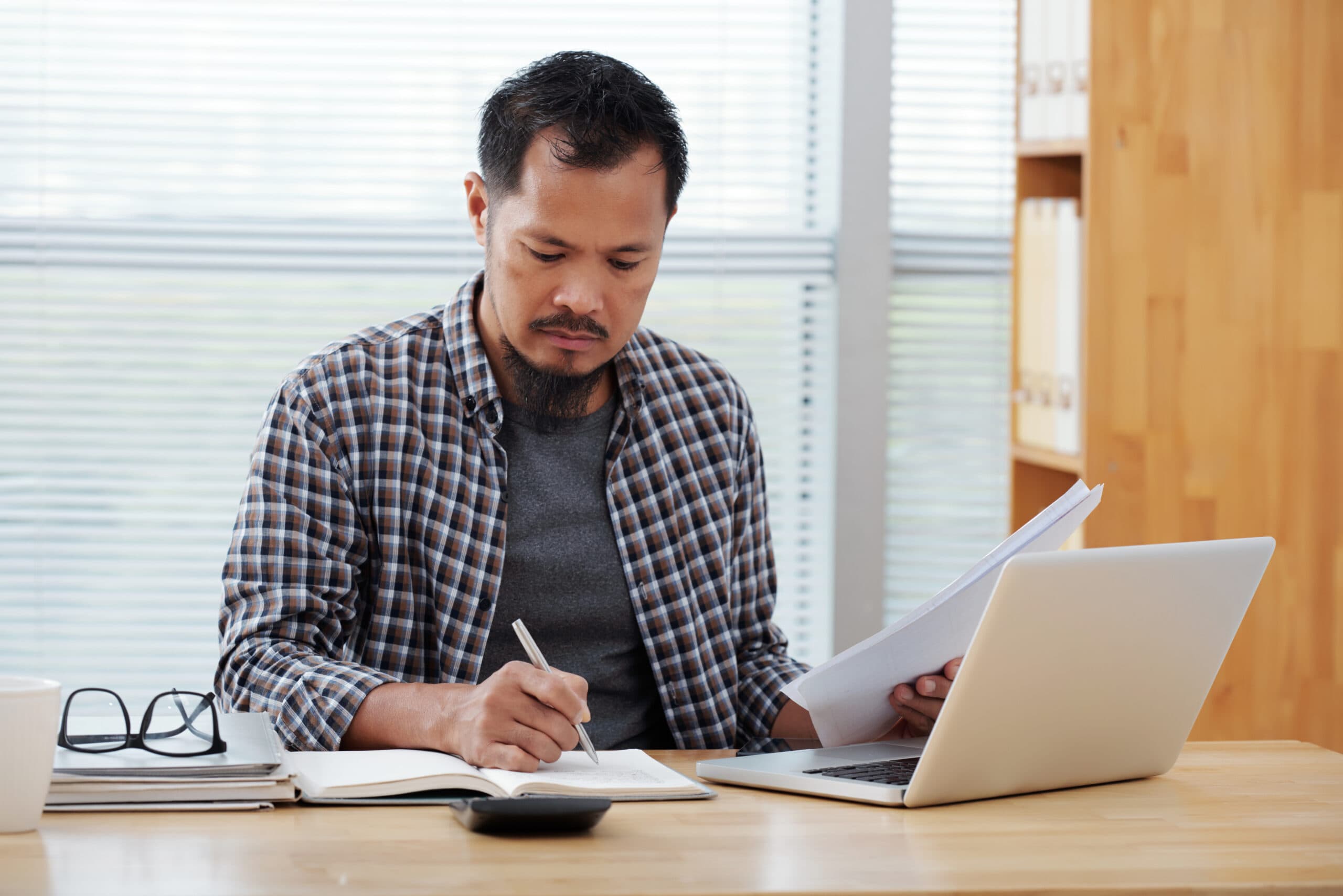 casually-dressed-asian-man-working-office-writing-notebook-holding-documents-scaled